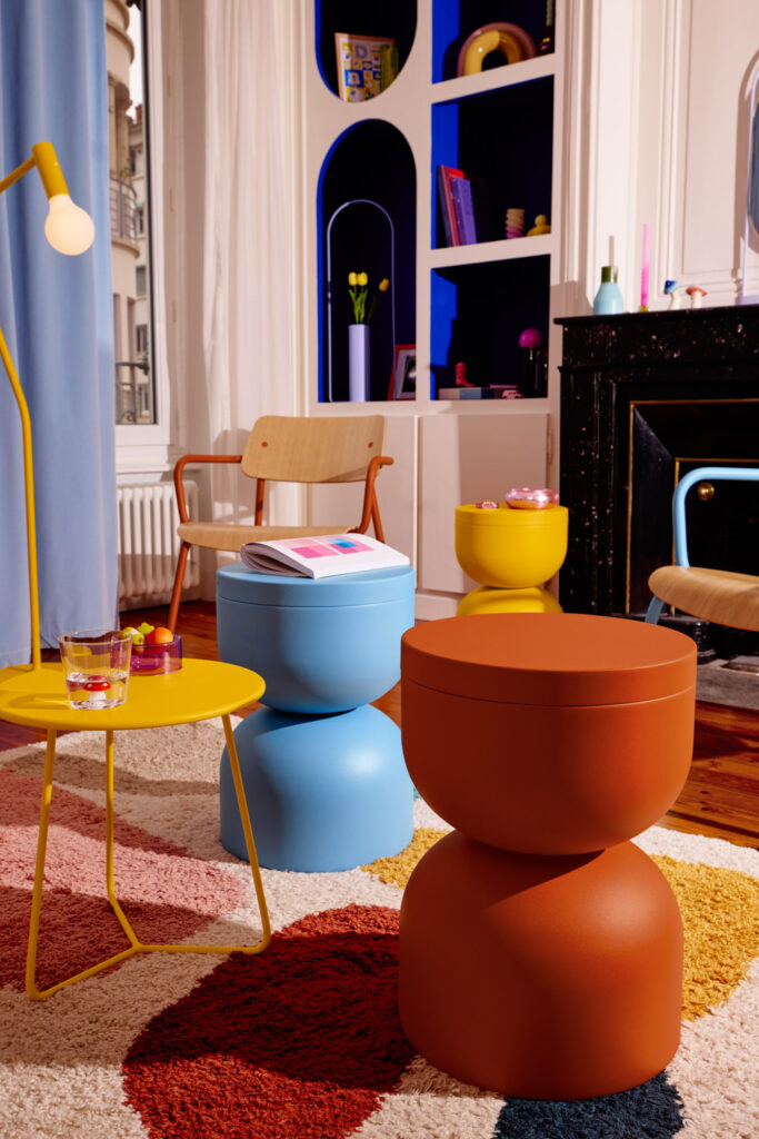 Un salon coloré avec un mobilier moderne, dont un tabouret Bleu Maya, des tabourets orange, une table d'appoint Piapolo jaune avec des boissons, une chaise et un tapis à motifs. Des étagères et des livres apportent du caractère à l'arrière-plan.