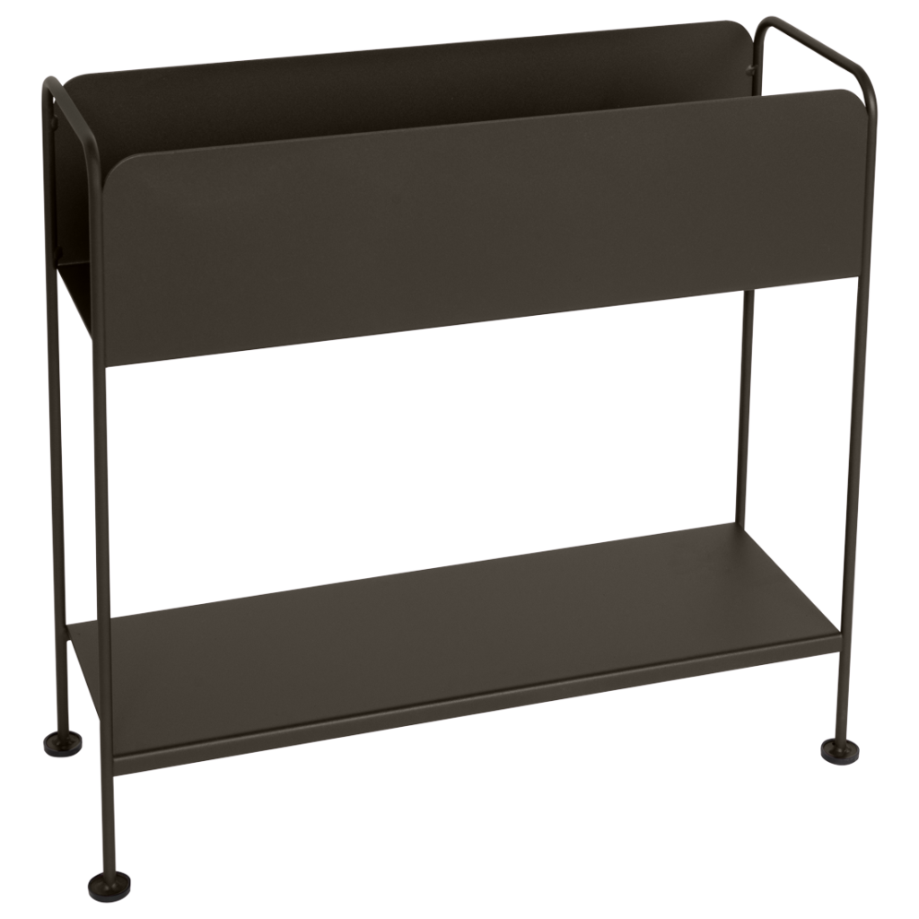 Un porte-revues en métal noir avec un compartiment supérieur et une étagère inférieure pour un rangement supplémentaire, posé sur quatre pieds à côté d'un cache-pot minimaliste, le tout sur un fond lavande uni.