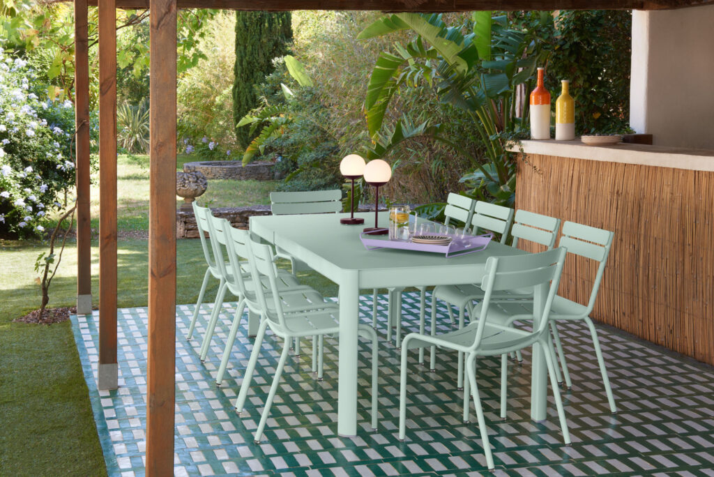 Une grande table à manger d'extérieur, comme la table extensible Ribambelle, est entourée de chaises vert menthe sous une pergola sur un patio carrelé, avec des plantes luxuriantes et un comptoir de bar Bleu Maya en arrière-plan.