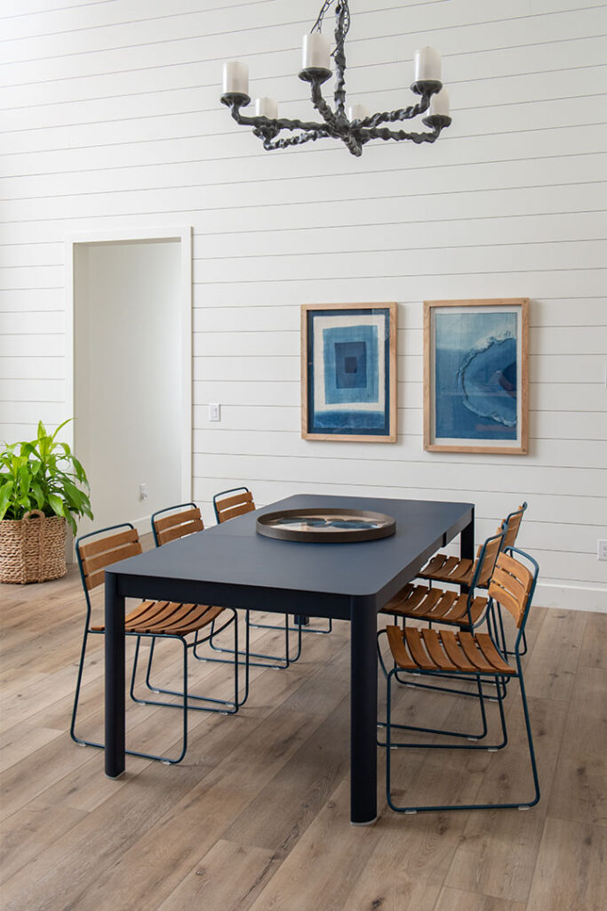 Une salle à manger moderne avec une table extensible Ribambelle noire, six chaises en bois, un lustre noir, deux tableaux abstraits bleus, une plante en pot et un parquet en bois clair.