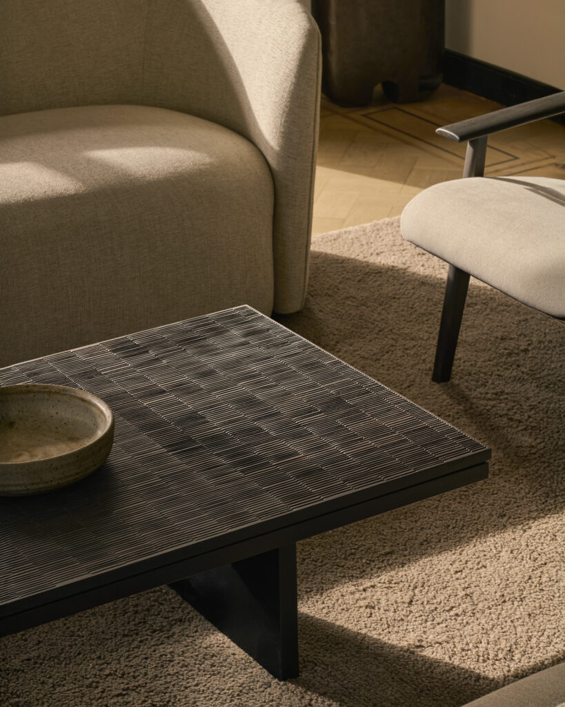 Un salon moderne avec un canapé beige, un fauteuil et une table basse Grooves en noir cassé comme pièce maîtresse, complétés par un bol en céramique et un tapis beige.