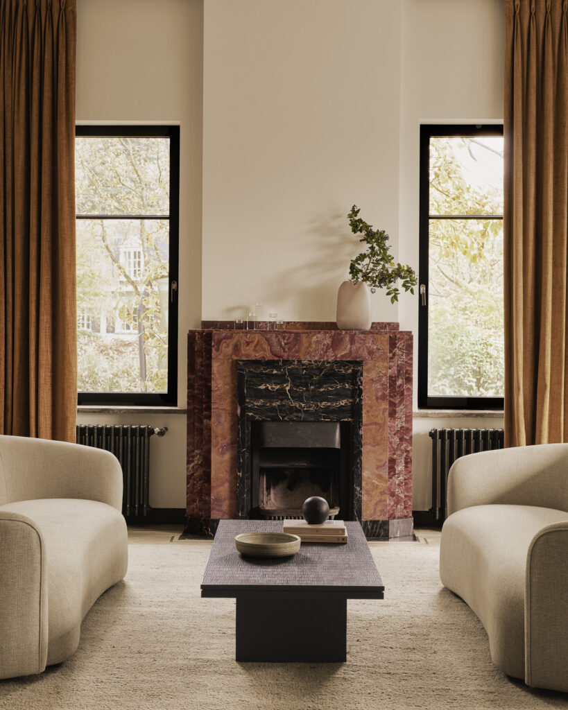 Salon contemporain avec canapés crème, table basse noir cassé, cheminée en marbre rouge et grandes fenêtres ornées de rideaux moutarde. Un vase orné de branches trône sur la cheminée.