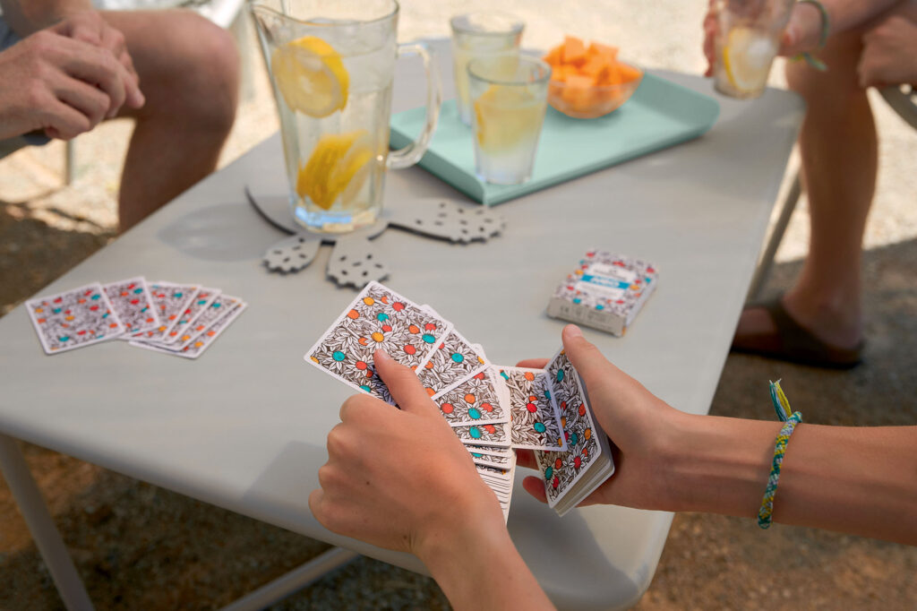 Deux personnes jouent aux cartes à une table basse sur la Croisette, avec des boissons fraîches et un plateau de fruits coupés. Au premier plan, l'une d'elles tient un éventail de cartes à jouer, idéal pour un après-midi de détente au bord de la mer bleu-maya.