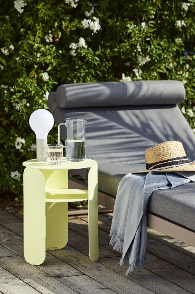 Une table d'appoint Bebop jaune avec un verre d'eau, un pichet et une lampe se trouve à côté d'une chaise longue d'extérieur rembourrée grise avec un chapeau de paille et une écharpe gris clair, posée sur une terrasse en bois.