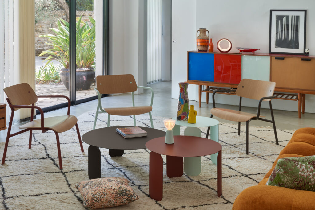 Salon moderne avec trois chaises minimalistes, deux tables basses rondes, un tapis à motifs, un meuble bas Bebop avec portes en verre coloré et une grande fenêtre laissant entrer la lumière naturelle.