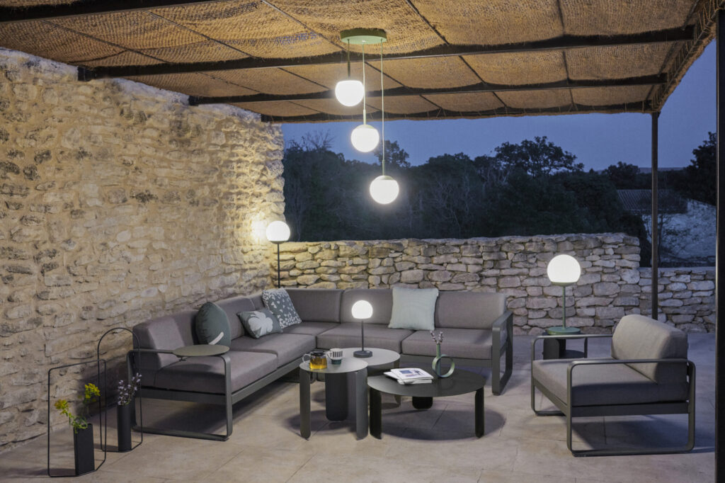 Espace extérieur avec canapé d'angle Bebop gris, fauteuil, table basse et suspensions rondes sous un auvent, entouré de murs en pierre à la tombée de la nuit. De subtiles touches d'Orange Confit réchauffent l'ambiance.
