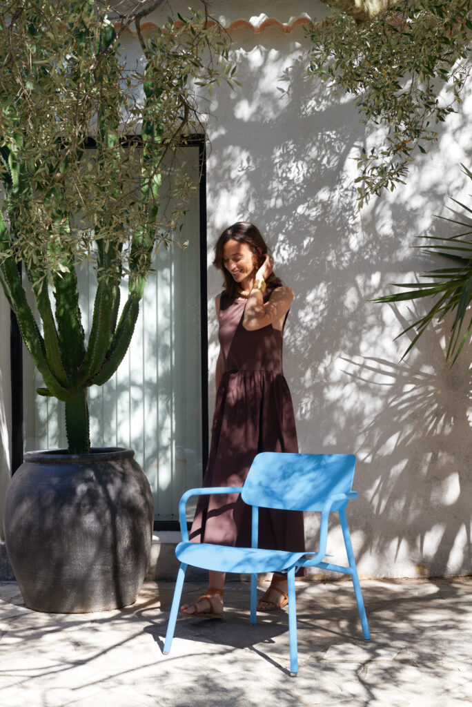 Une femme vêtue d'une robe marron sans manches se tient dehors près d'un grand cactus en pot, avec une chaise Fauteuil Lounge Studie devant elle tandis que la lumière du soleil projette des ombres sur le mur.