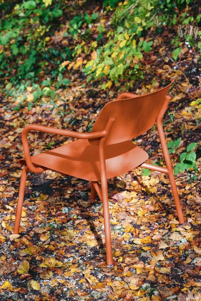 Un fauteuil Lounge Studie orange est placé à l'extérieur sur des feuilles mortes, entouré d'un feuillage vert luxuriant.