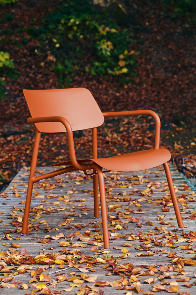 Le Fauteuil Lounge Studie, une chaise orange moderne, repose sur une surface en bois parsemée de feuilles d'automne, sur fond naturel légèrement flou.