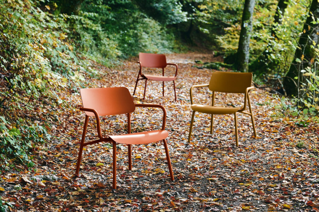 Trois chaises colorées Fauteuil Lounge Studie sont posées sur un chemin forestier parsemé de feuilles, entouré d'un feuillage vert luxuriant et d'arbres.