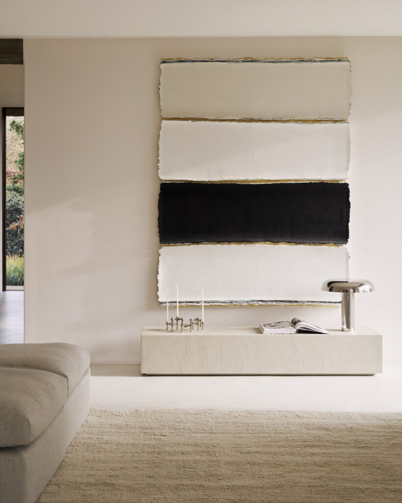 Salon minimaliste avec un grand tableau abstrait, des murs beiges, un canapé et un tapis clairs, et l'étagère au sol Elements présentant des bougies, un livre et une lampe métallique.