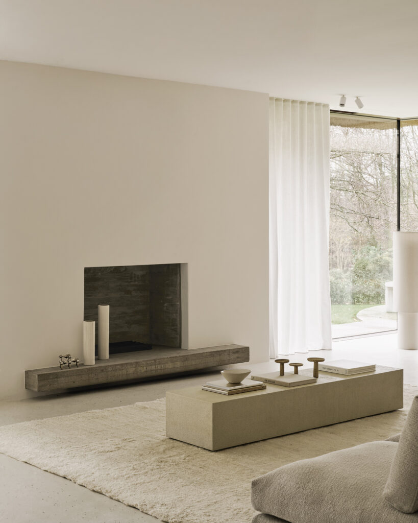 Un salon minimaliste dans des tons neutres comprend une cheminée, une table basse moderne avec décor, un tapis, une étagère au sol Elements, et de grandes fenêtres avec des rideaux diaphanes donnant sur un jardin.