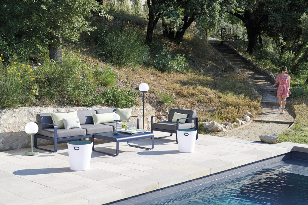 Espace salon extérieur moderne avec canapés Bellevie gris, table basse et deux poubelles blanches à côté d'une piscine ; une femme en robe rose marche dans un chemin de jardin en arrière-plan.