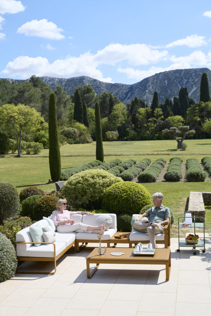 Deux personnes se détendent sur des canapés d'extérieur Bellevie sur une terrasse, avec des boissons posées sur une table basse élégante, surplombant un jardin paysager et des montagnes sous un ciel partiellement nuageux.