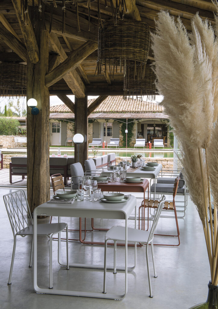 Espace repas extérieur avec table moderne Bellevie 74 x 80 cm et chaises ton tonka sous une pergola en bois, dressée avec assiettes et verres, et décorée d'herbe de la pampa.