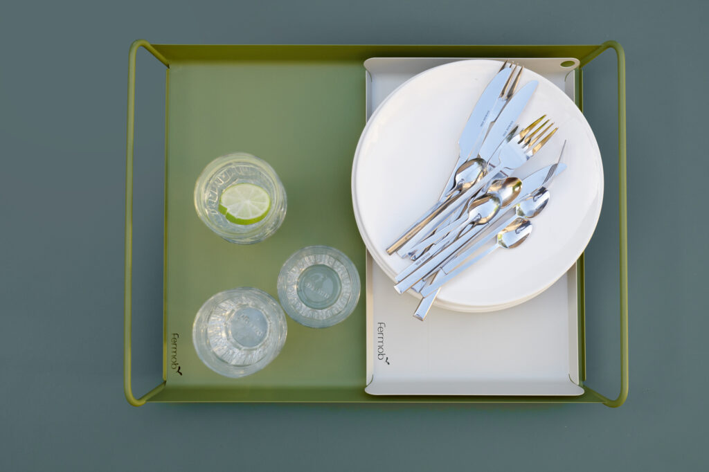 Deux plateaux verts Plateau Alto (53 x 38 cm) contiennent deux verres d'eau avec du citron vert, deux verres vides et une pile d'assiettes blanches avec des couverts dessus, le tout disposé soigneusement dans un style bleu acapulco rafraîchissant.