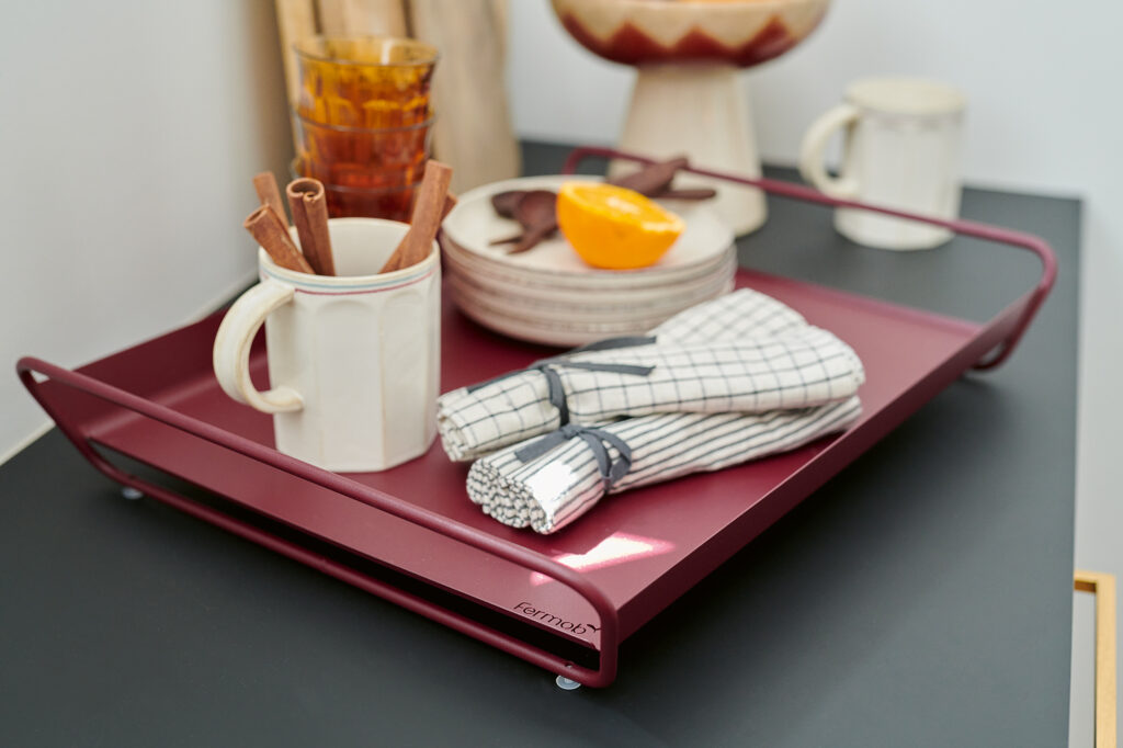 Un plateau marron Plateau Alto, 53 x 38 cm, contient une tasse avec des bâtons de cannelle, une pile de bols, des serviettes, un verre, un quartier d'orange et une tasse sur une surface sombre.