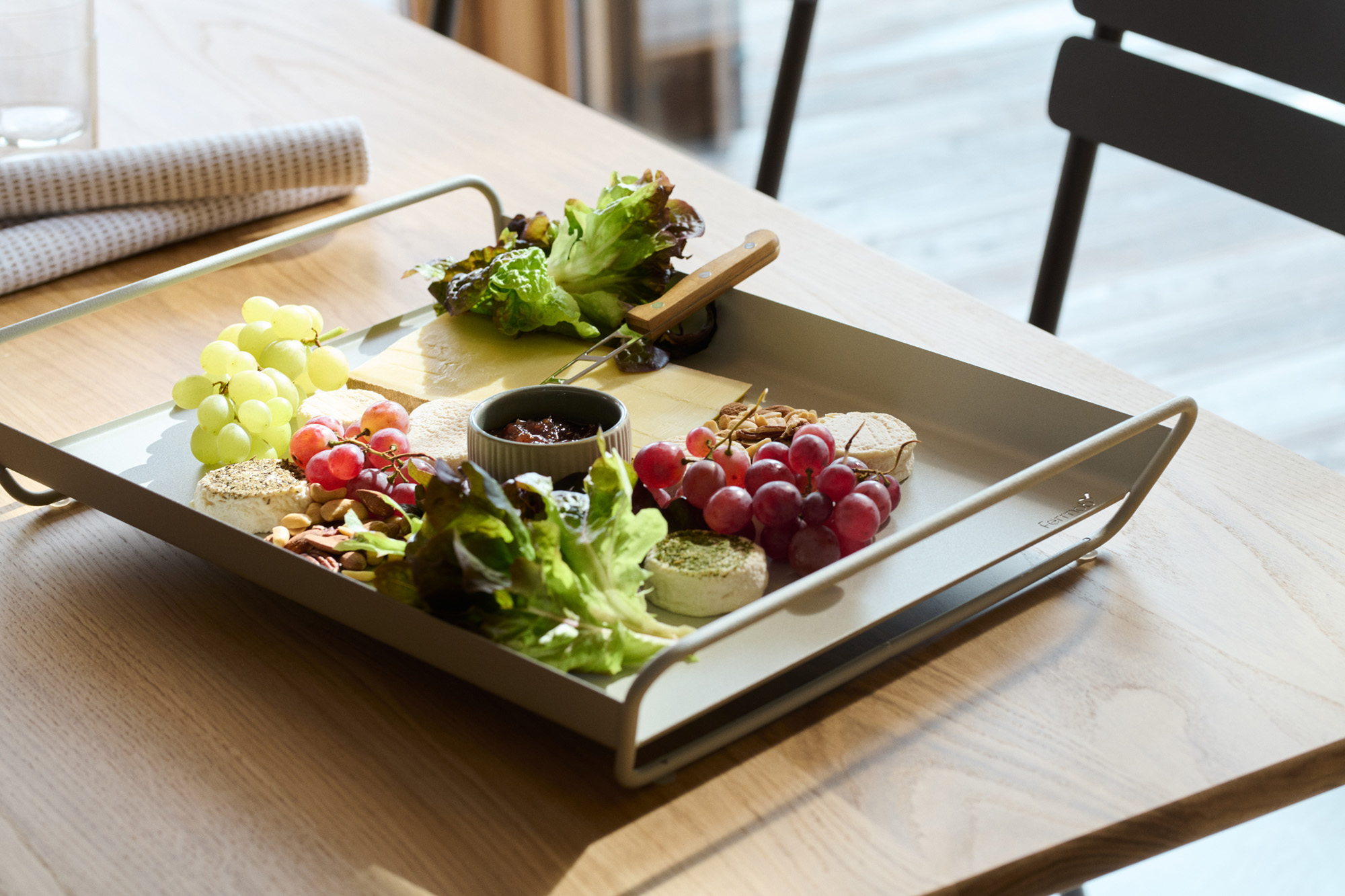 Un Plateau Alto de 53 x 38 cm repose sur une table en bois, agrémenté d'un assortiment de fromages, de crackers, de raisins verts et rouges, de légumes verts et d'une petite coupelle de confiture. Une part de Bleu Acapulco apporte une touche gourmande.