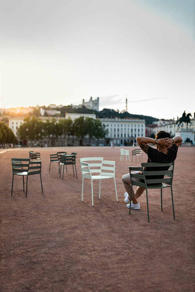 Un homme est assis sur une chaise Fauteuil Rythmic, les mains derrière la tête, sur une place ouverte avec des chaises vides éparpillées, des bâtiments et des arbres en arrière-plan au coucher du soleil, le tout baigné d'une douce lumière gris orage.