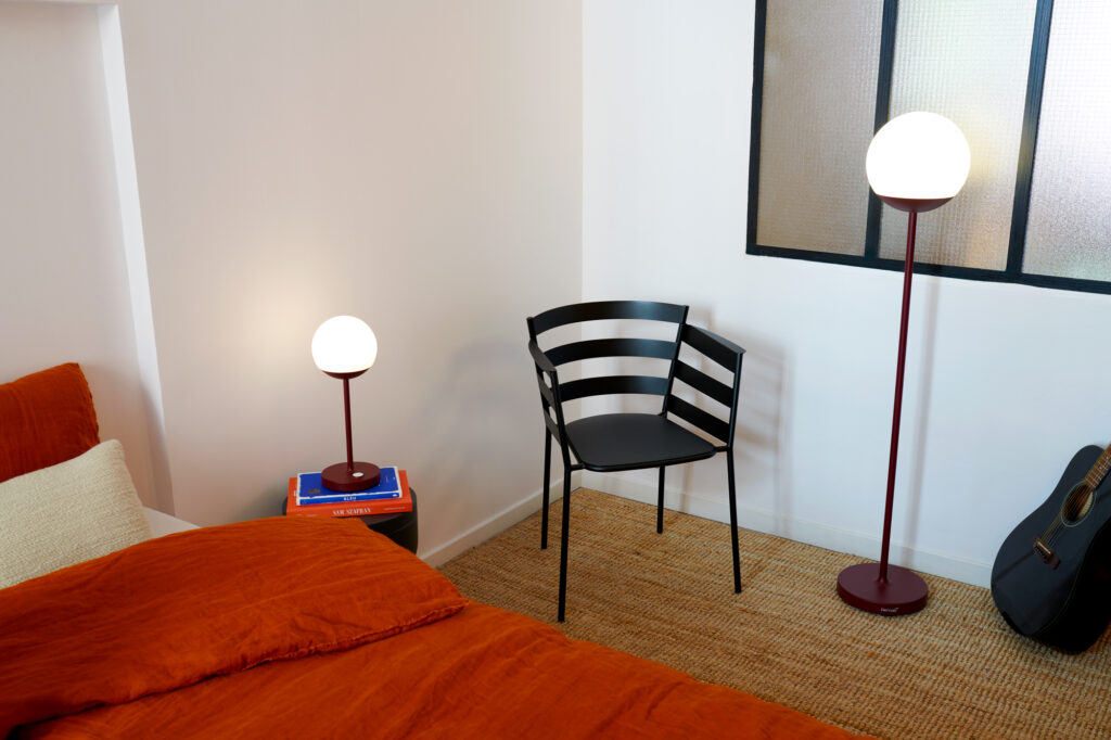 Une chambre avec une literie orange, un Fauteuil Rythmic en Gris orage, une petite lampe sur une table de chevet avec des livres, un lampadaire, une guitare et une fenêtre avec du verre texturé.