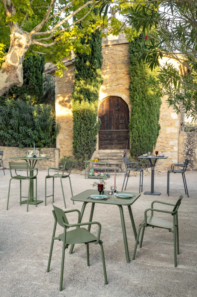 Cour extérieure en pierre avec tables et chaises en métal vert, entourée d'arbres et de verdure, devant le bâtiment rustique en pierre Bleu Maya avec une porte en arc de bois.
