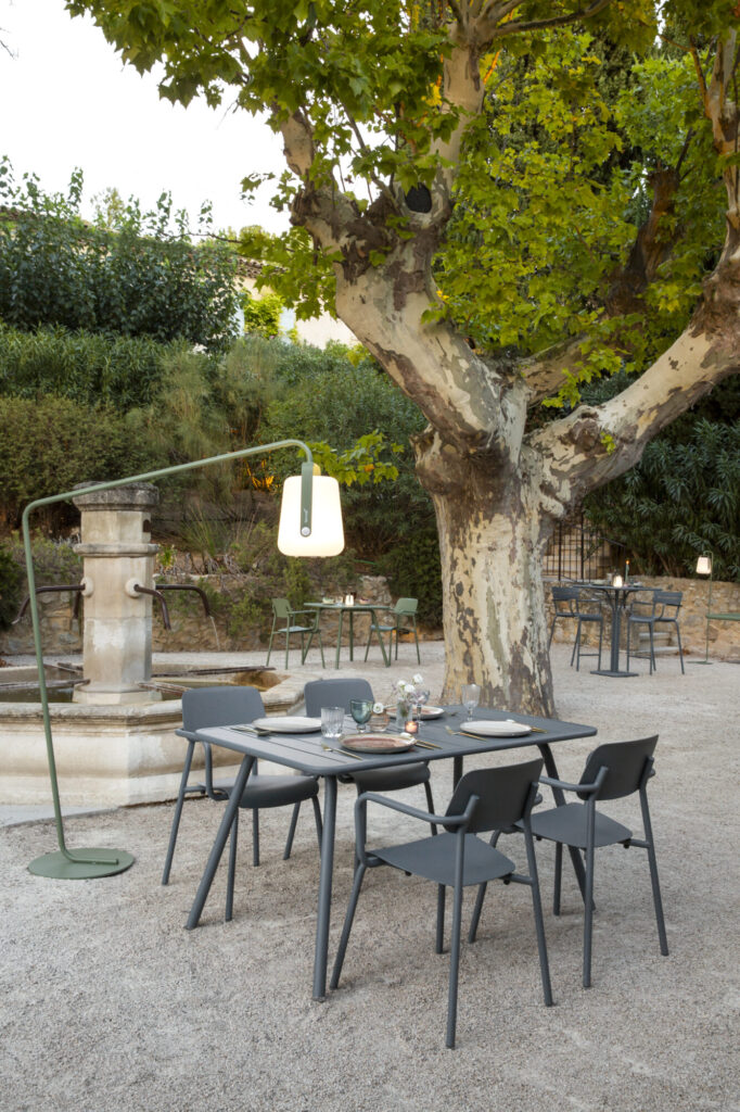 Espace repas extérieur avec tables et chaises grises sur gravier, installé sous un grand arbre avec un lampadaire et une fontaine en pierre Bleu Maya en arrière-plan.