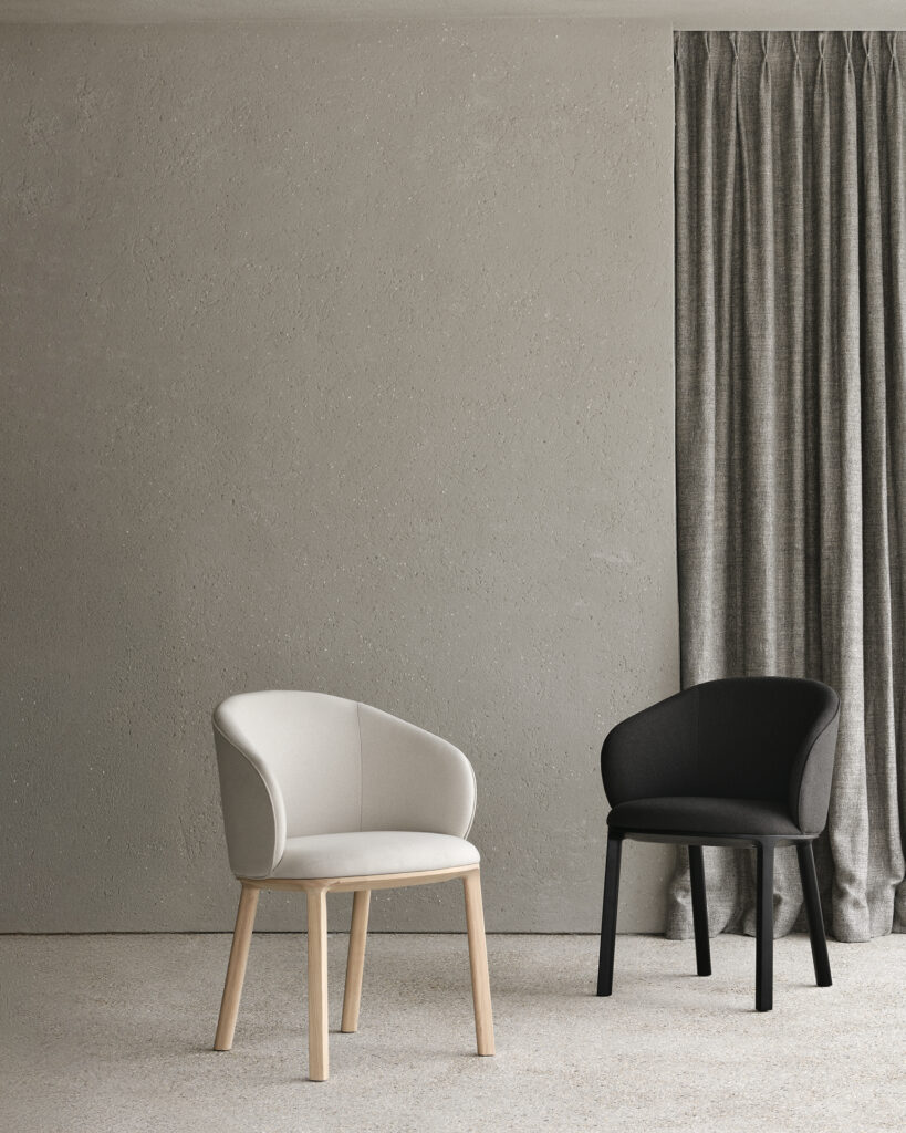 Une chaise de salle à manger Unda beige et une chaise noire sont disposées côte à côte dans une pièce minimaliste aux murs gris, avec un rideau et de la moquette. Des touches de chêne apportent de la chaleur à la palette neutre.
