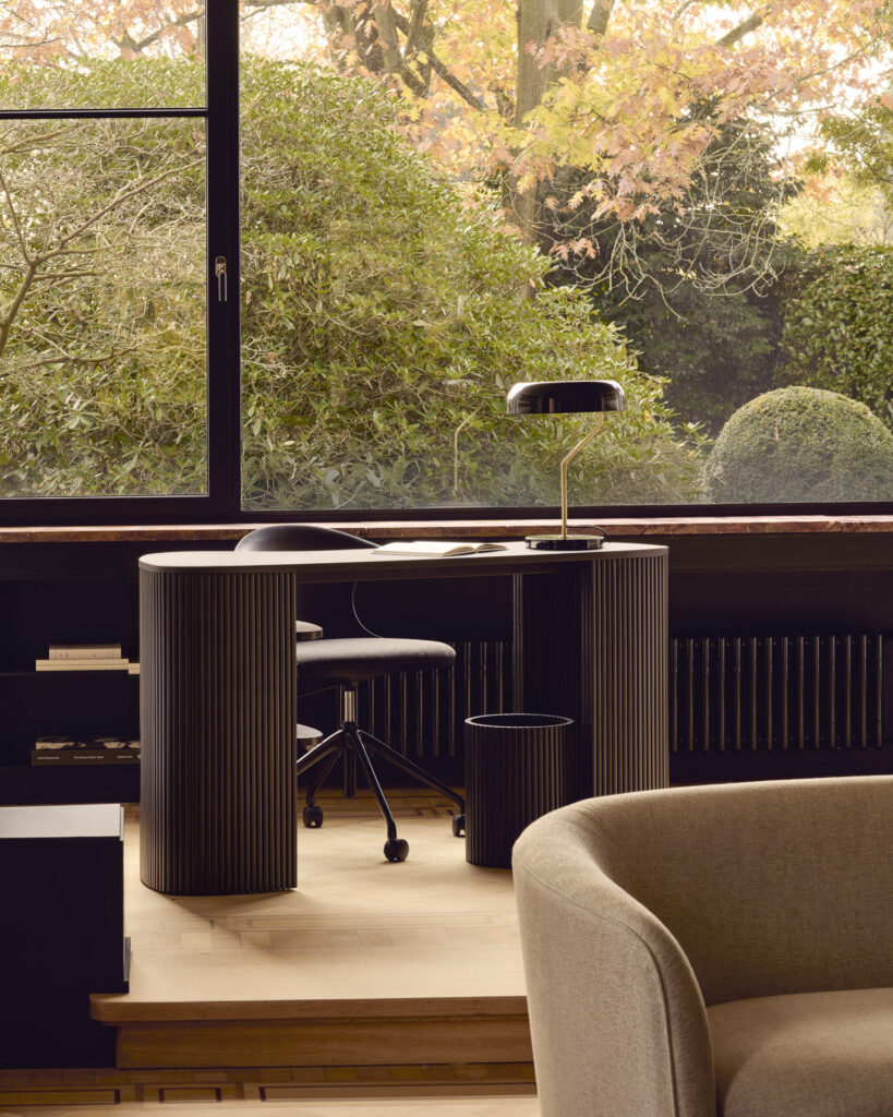 Un élégant bureau Roller Max noir est associé à une lampe et une chaise près d'une grande fenêtre, offrant une vue sur la verdure luxuriante et le feuillage aux couleurs d'automne du jardin.