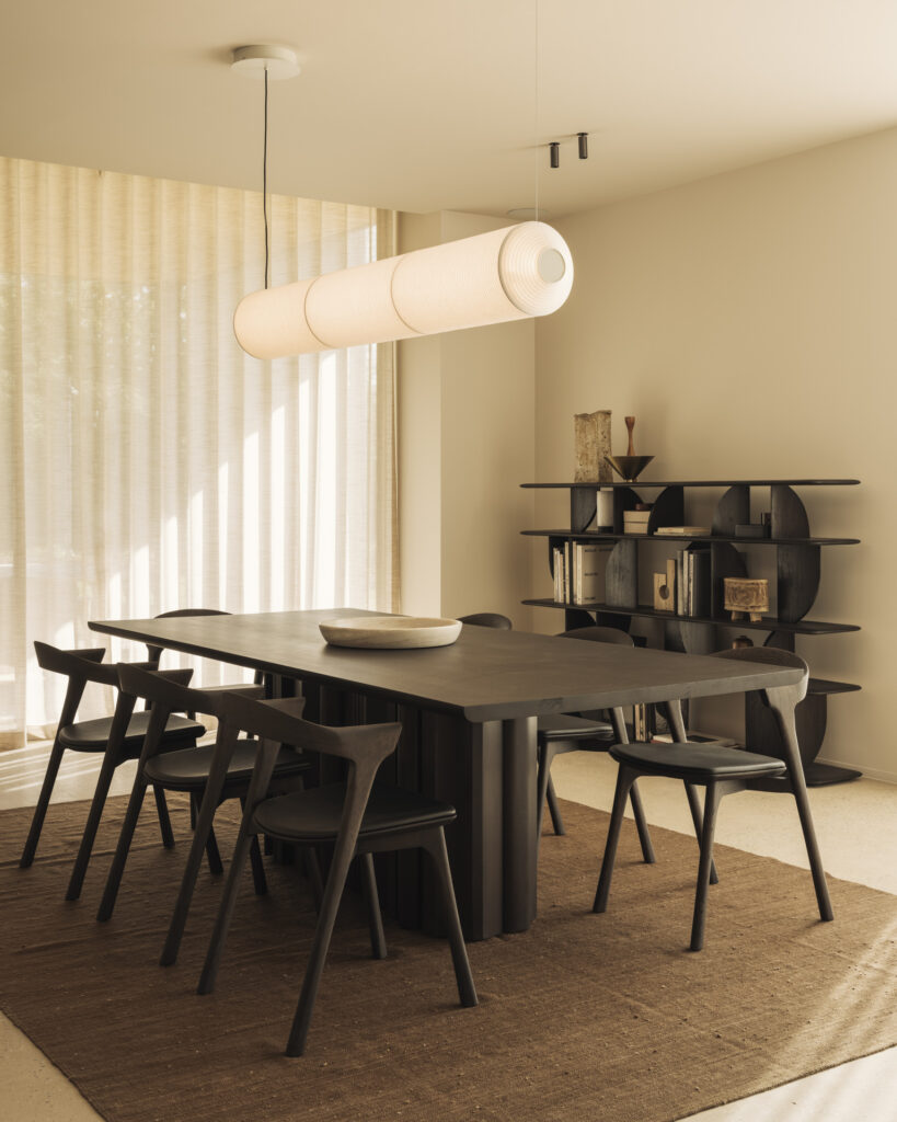 Une salle à manger minimaliste présente la table à manger Sequence et ses six chaises, associées à un tapis rectangulaire, une lampe suspendue moderne, des étagères ouvertes, des murs et des rideaux de couleur claire.