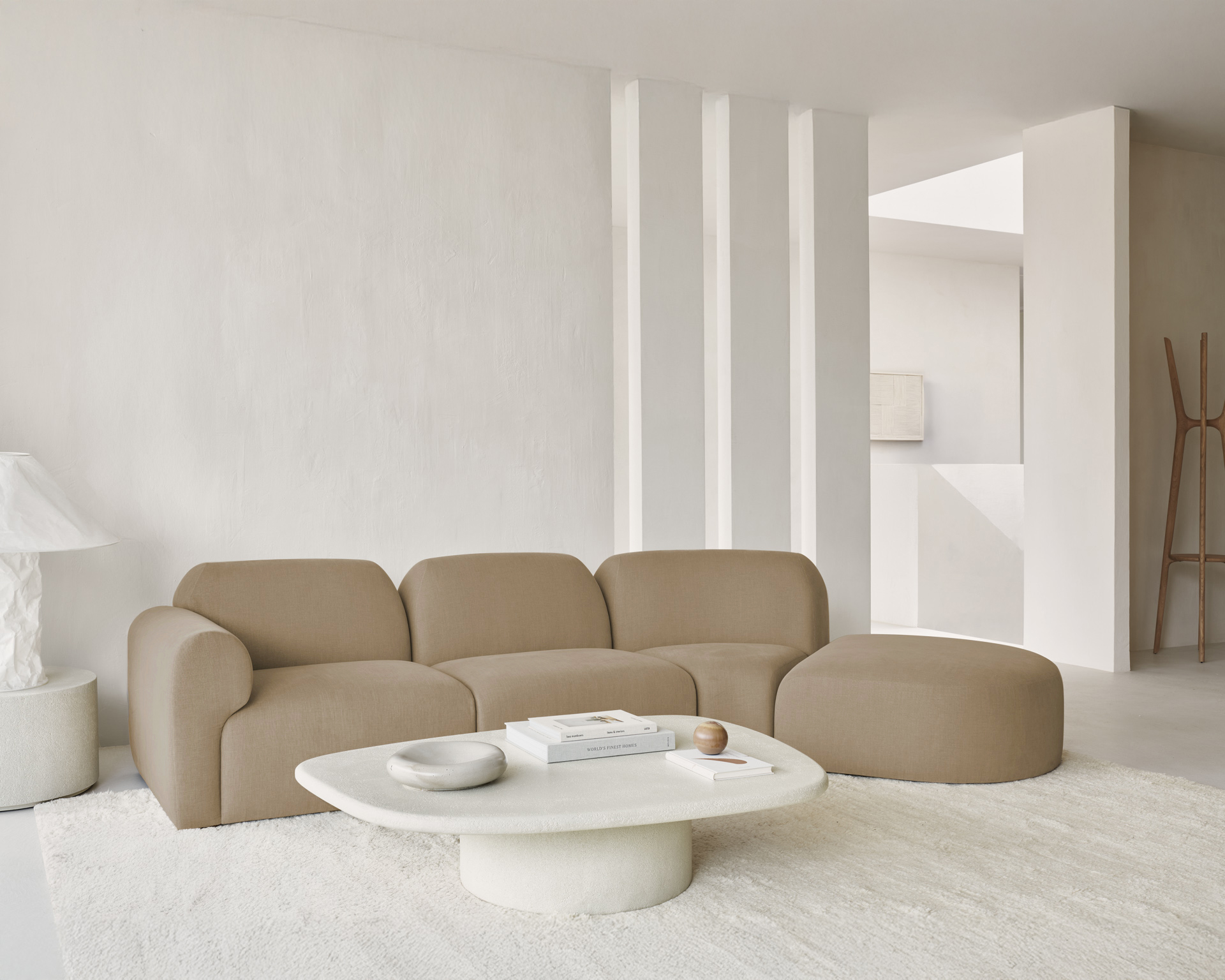Salon minimaliste avec le canapé beige courbé Canapé Bulky, une table basse ovale blanche, une lampe blanche et des murs crème, mis en valeur par une douce lumière naturelle et un décor neutre.