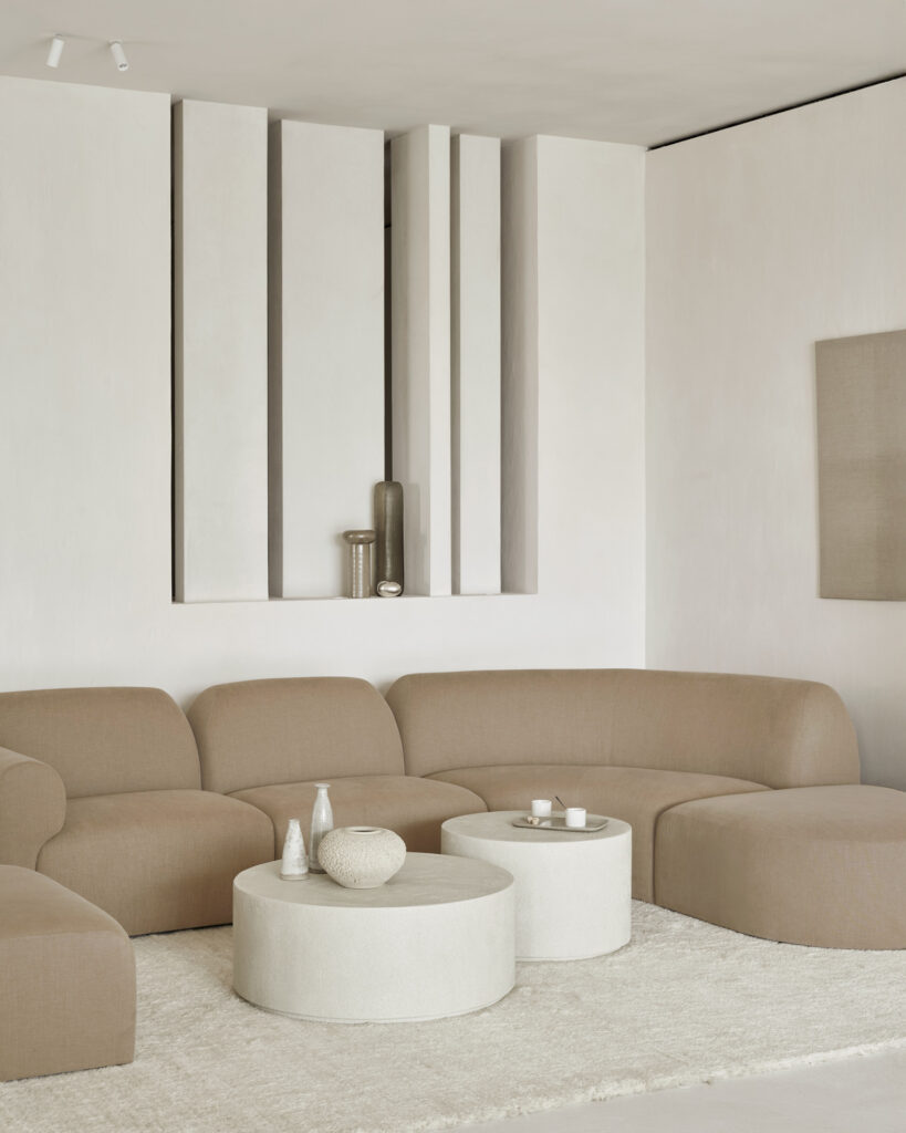 Un salon moderne comprend un canapé beige incurvé Canapé Bulky, deux tables basses blanches rondes, des accents en céramique et un élément mural texturé aux tons neutres.