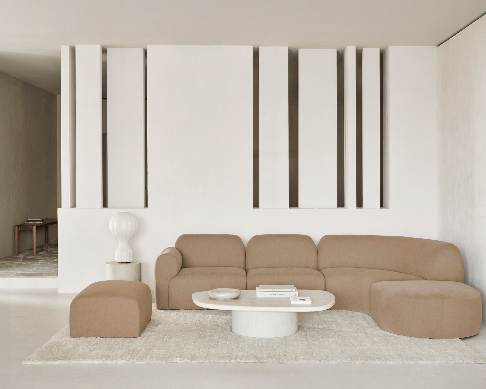 Un salon moderne comprenant le canapé sectionnel et le pouf Canapé Bulky beige, une table basse blanche, une lampe blanche et des livres décoratifs placés sur un tapis de couleur claire.