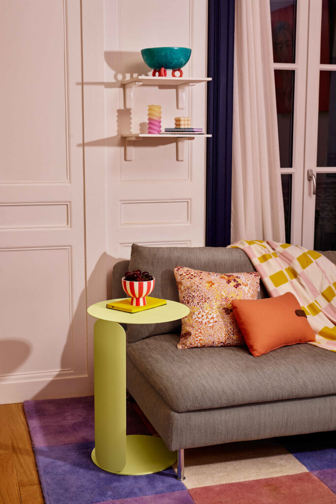 Un salon moderne avec un canapé gris, des coussins colorés, une table déportée Bebop en vert surmontée d'un bol de cerises, un tapis violet et jaune, et des étagères murales blanches présentant des objets décoratifs.