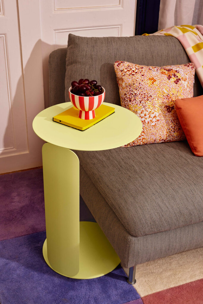 Une petite Table déportée Bebop vert clair est assise à côté d'un canapé gris, tenant un bol de raisins rayé et un livre jaune fermé. Des coussins à motifs et un tapis coloré sont également visibles.