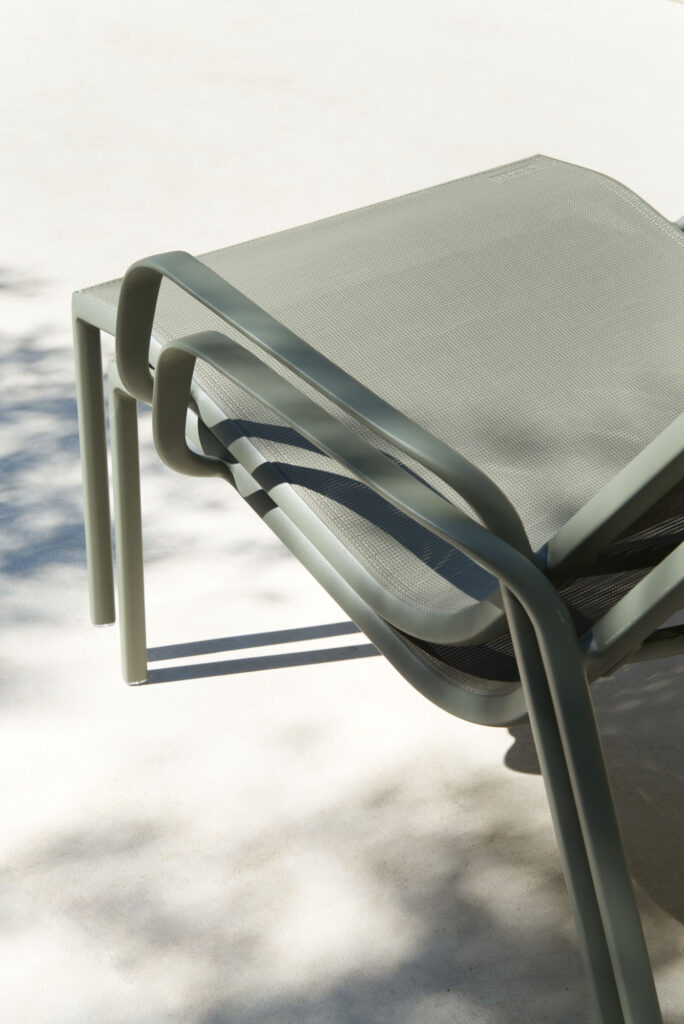 Deux chaises Fauteuil Lounge Cadiz empilées, en métal et maille vert olive, reposent sur une surface claire, partiellement à l'ombre.