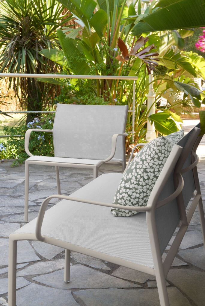 Le duo Banquette Cadiz, deux chaises de patio en métal blanc, dont l'une avec un coussin à motifs, repose sur une terrasse en pierre encadrée de plantes tropicales luxuriantes.