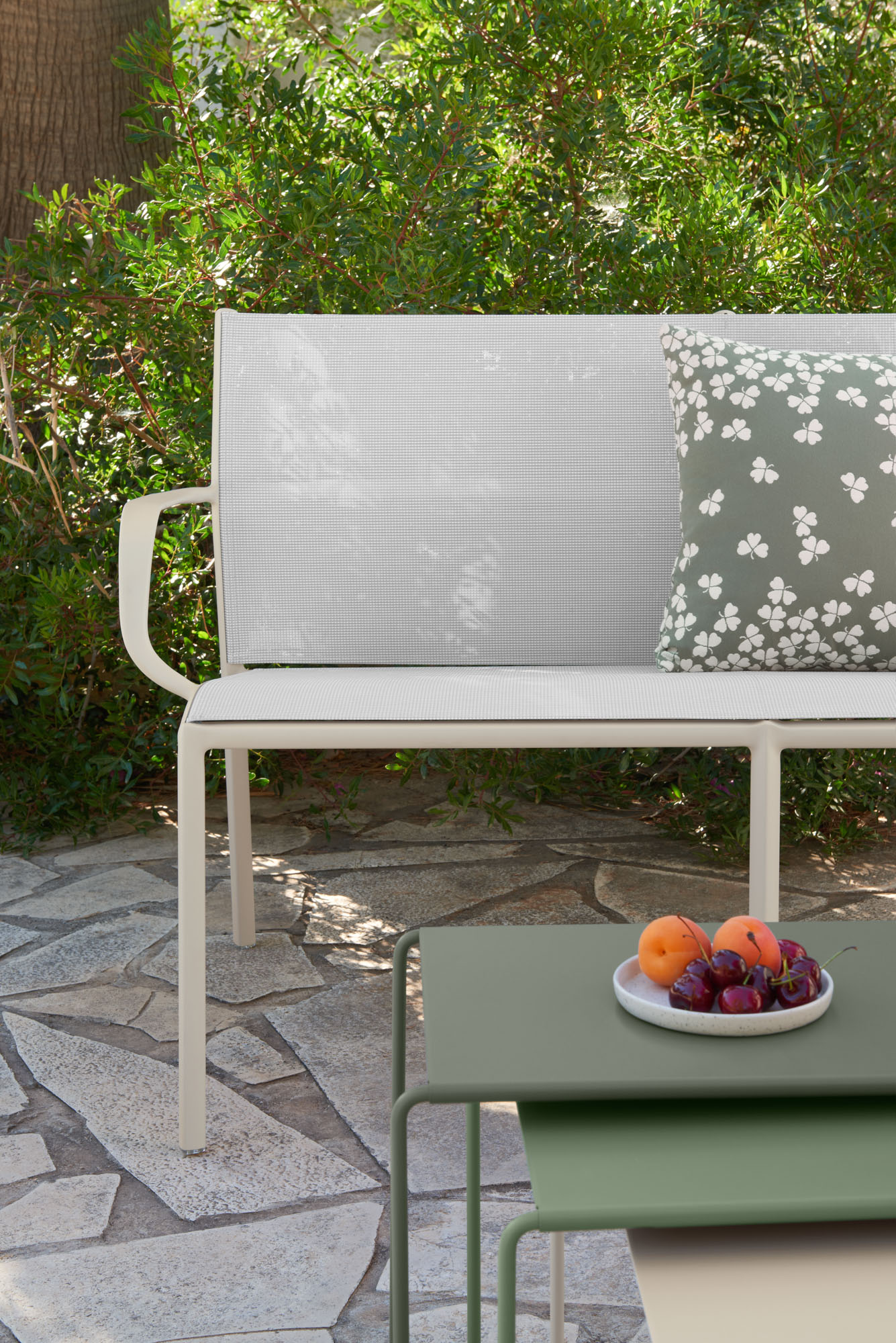 Le duo de banquettes Cadiz présente un banc d'extérieur blanc avec un coussin floral vert sur un patio en pierre, associé à deux tables emboîtées contenant des fruits. De la verdure luxuriante complète ce décor accueillant.