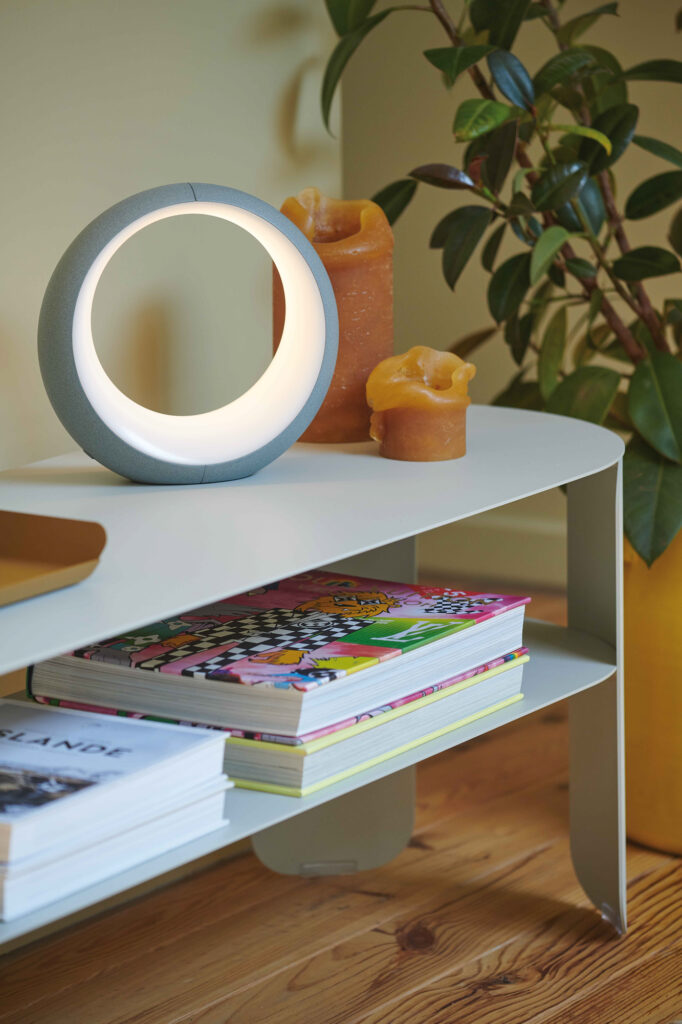 Une lampe circulaire moderne, trois bougies ambrées et des magazines colorés sont posés sur la Console basse Bebop à côté d'une plante en pot, apportant une touche Bebop élégante à l'espace.