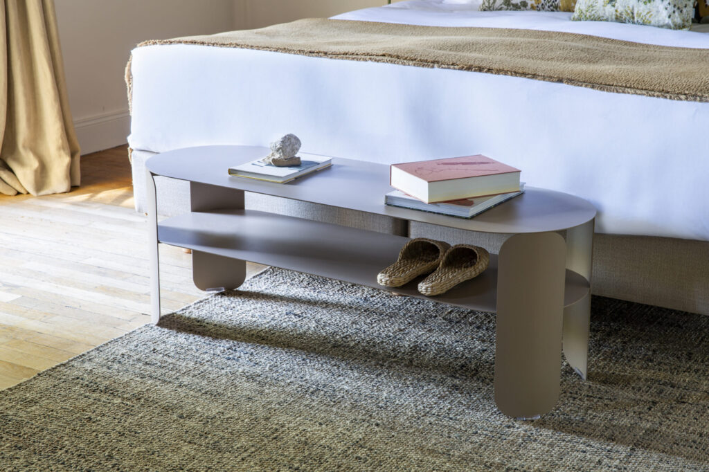 Un banc moderne, ovale et gris, inspiré de la console basse Bebop et doté de deux étagères, trône au pied d'un lit et accueille des livres, des objets de décoration et des pantoufles tissées sur un tapis texturé dans une chambre lumineuse.