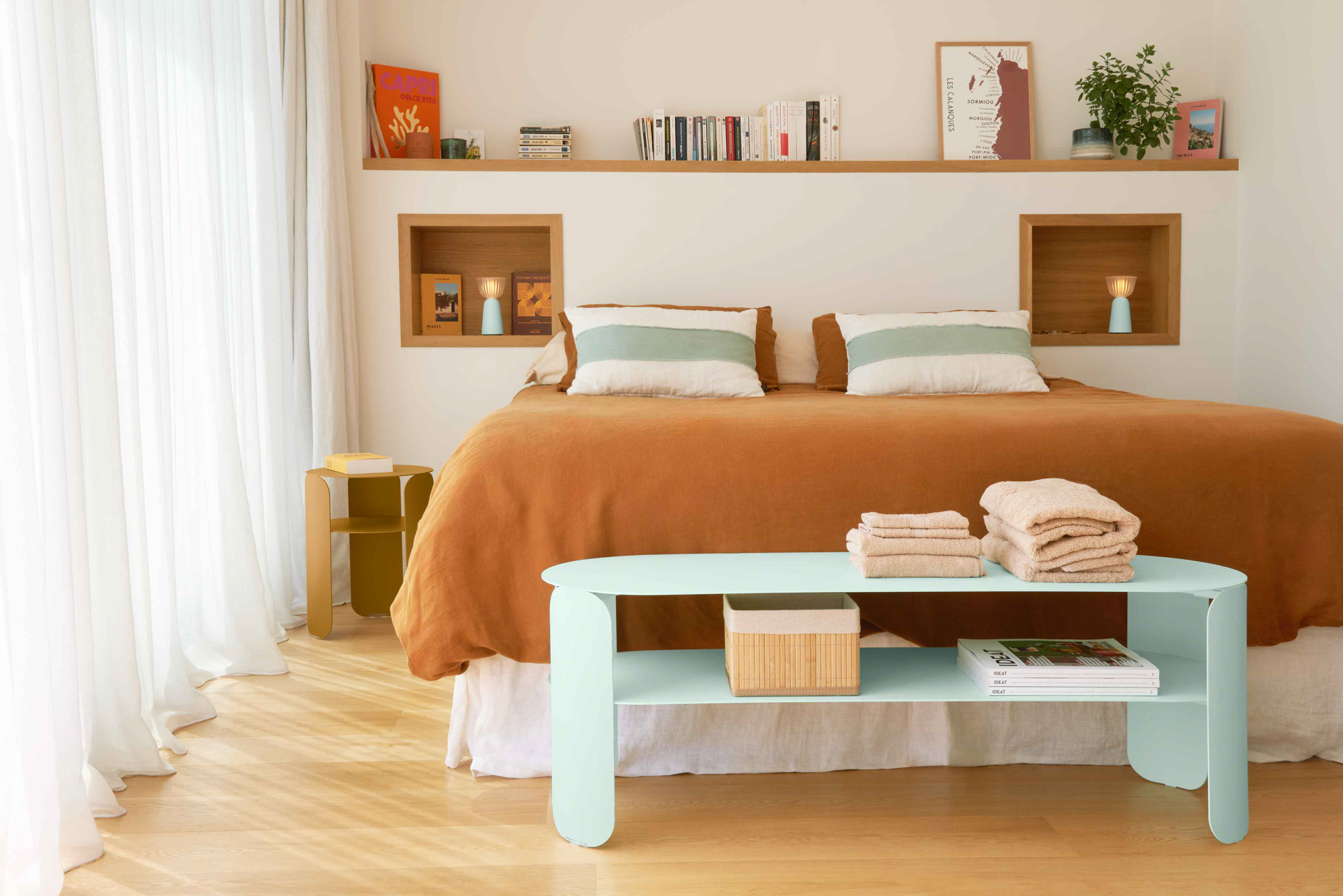 Une chambre à coucher moderne présente un couvre-lit marron, deux oreillers et une Console basse Bebop bleu clair avec des serviettes pliées, tandis qu'une étagère flottante au-dessus du lit présente des livres et de la décoration.