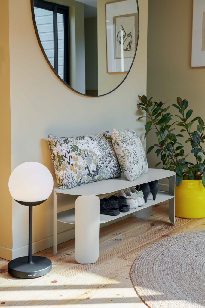 Une petite entrée avec un banc à coussin fleuri, des chaussures sur l'étagère inférieure de la Console basse Bebop, un miroir mural rond, une plante en pot dans un pot jaune et un lampadaire moderne créent une ambiance accueillante inspirée de Bebop.