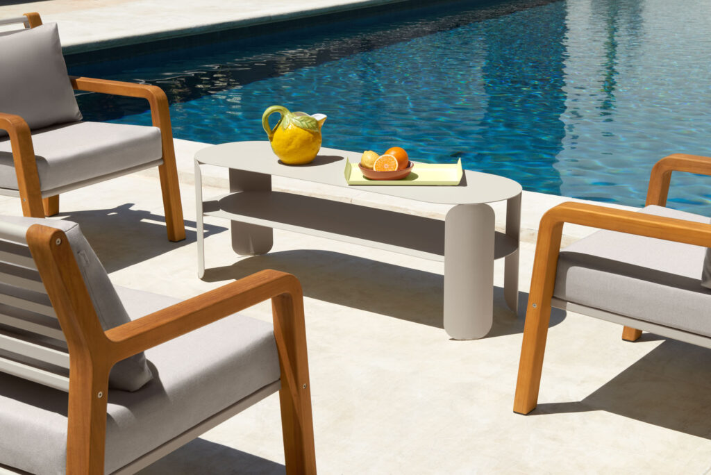 La Console basse Bebop, associée à trois chaises en bois rembourrées et à une table blanche garnie de fruits et d'un pichet, est installée au bord de la piscine à la lumière du jour, créant ainsi une atmosphère de détente en plein air.