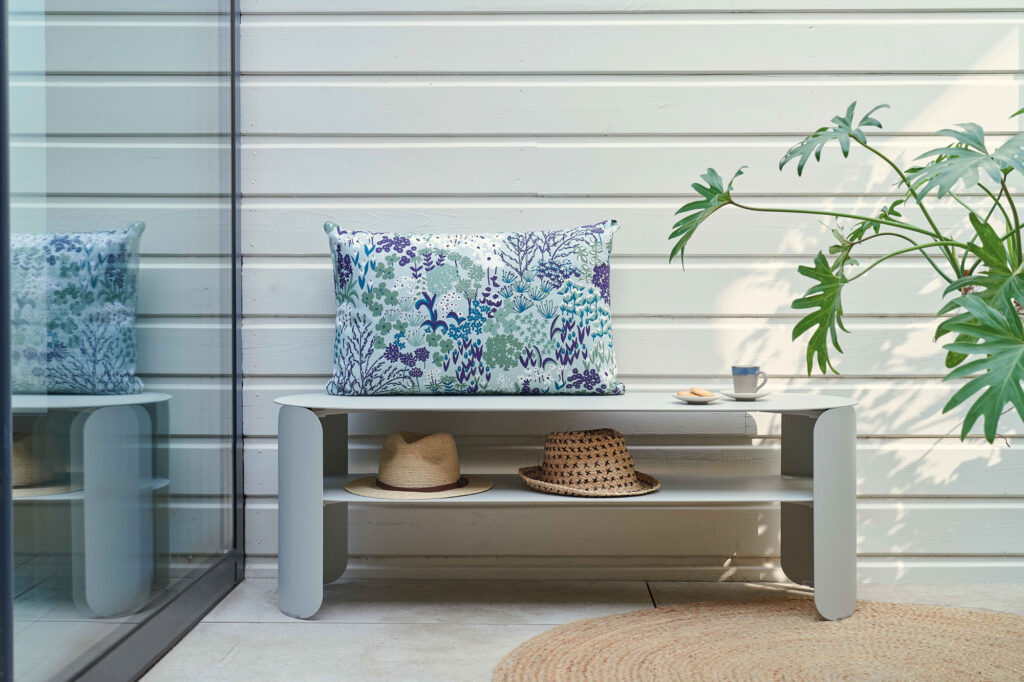 La console basse Bebop - un banc gris avec un coussin fleuri, des chapeaux en dessous, une tasse et un livre à côté d'une plante verte - ajoute du charme à votre espace contre un mur lambrissé blanc.