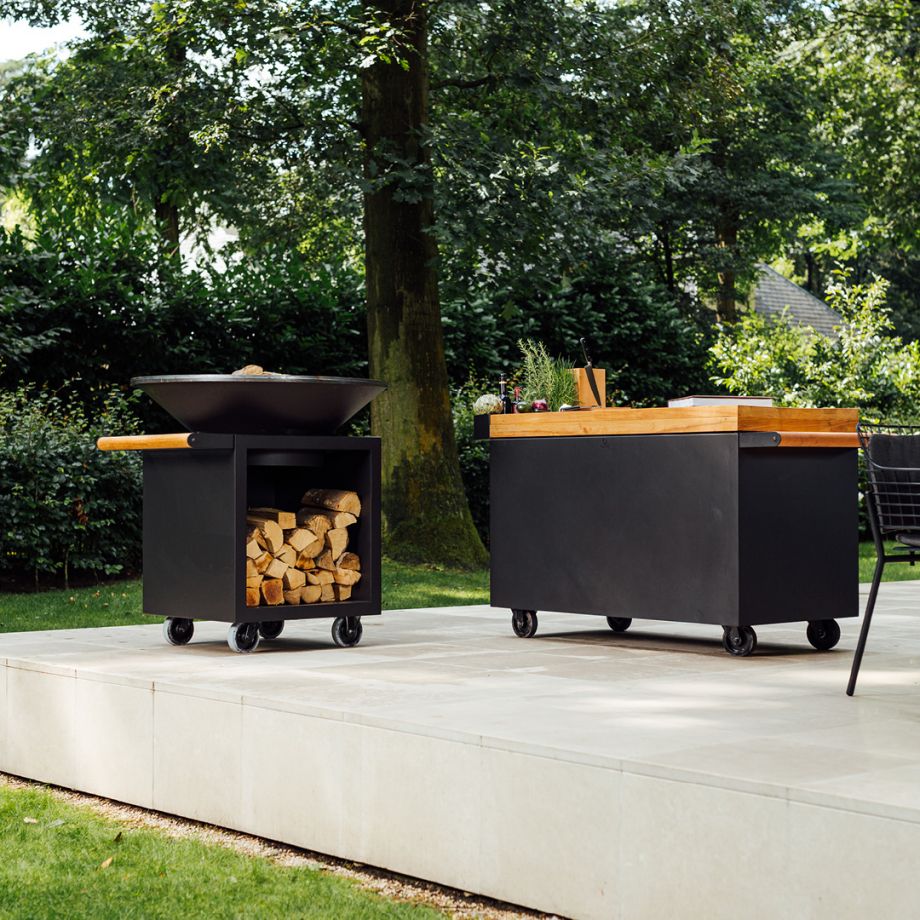 Deux chariots de cuisine d'extérieur noirs sur roues, l'un doté d'un rangement en bois avec un bol à feu sur le dessus, l'autre d'un comptoir en bois, sont situés sur un patio dans un jardin. Parfait pour une cuisine d'extérieur élégante avec une touche Ofyr PRO.