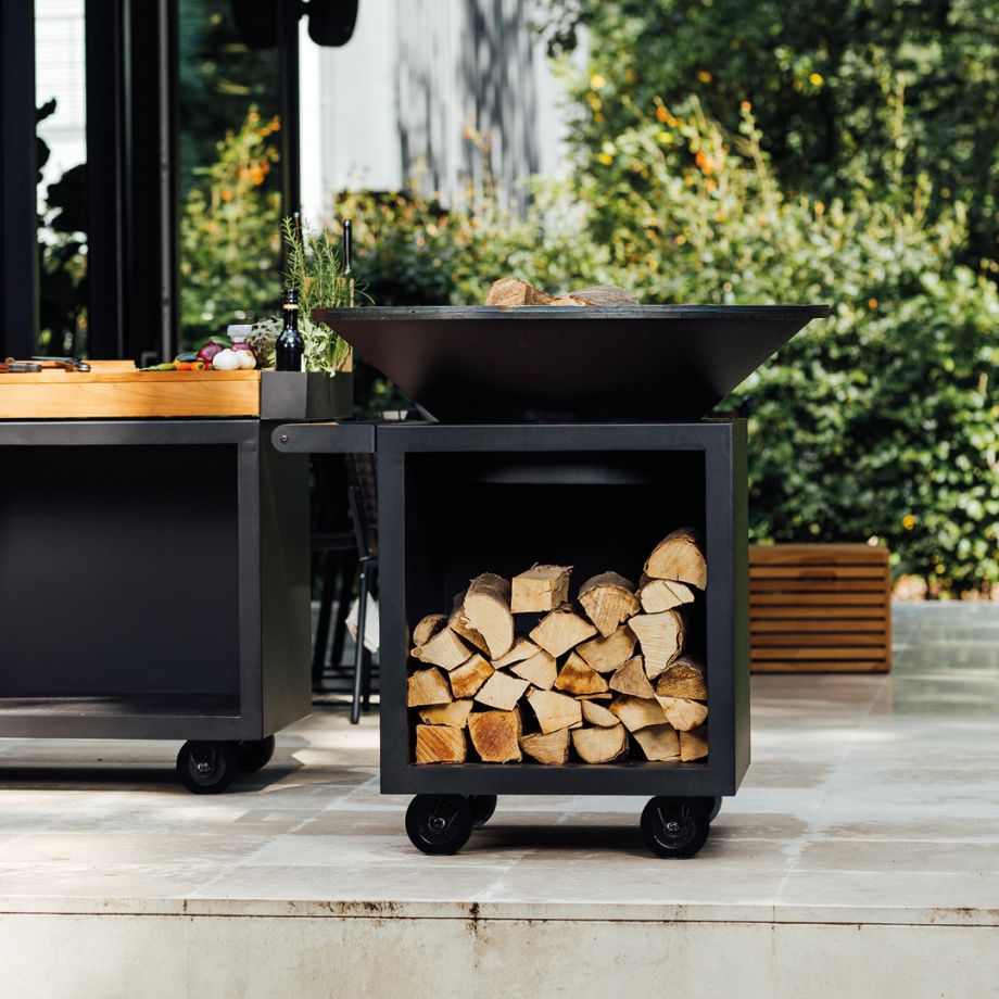 Un foyer d'extérieur sur roues en métal noir Ofyr PRO, doté d'un compartiment de rangement pour le bois de chauffage fendu, se dresse sur un patio carrelé à côté d'un comptoir en bois.