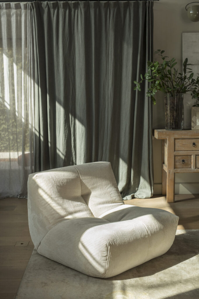 La chaise longue crème Cozy S - 1 place repose sur un tapis beige, à côté de rideaux longs et d'une table d'appoint en bois avec une plante en pot. La lumière du soleil passe à travers la fenêtre, ce qui ajoute à son style moderne et accueillant.
