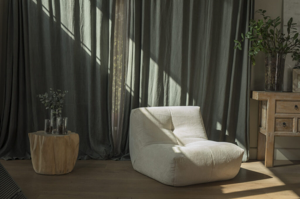 Une pièce ensoleillée présente la chaise longue Cozy S - 1 place blanc doux et une petite table d'appoint en bois avec des vases, placée près d'une fenêtre avec des rideaux verts et des plantes d'intérieur.