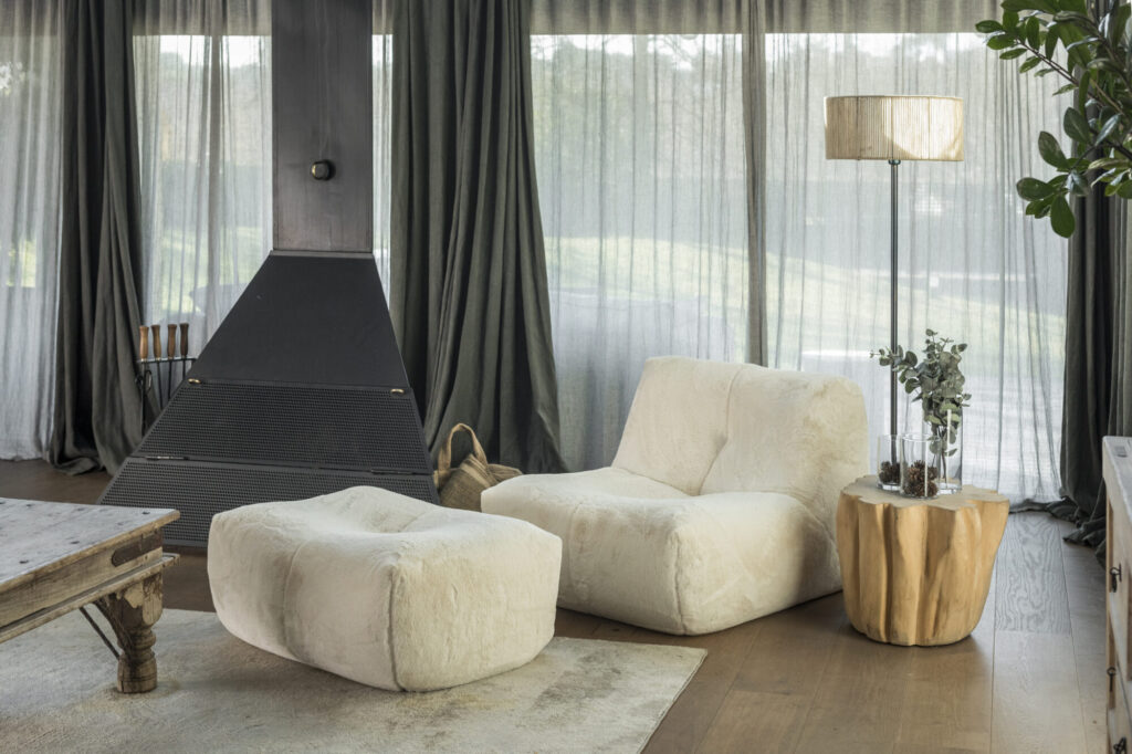 Un salon moderne avec le Cozy S - 1 place CZ1 + Pouf CZ23, une cheminée noire, un fauteuil beige, une table d'appoint en bois avec lampe et vase, et de grandes fenêtres drapées de rideaux diaphanes.