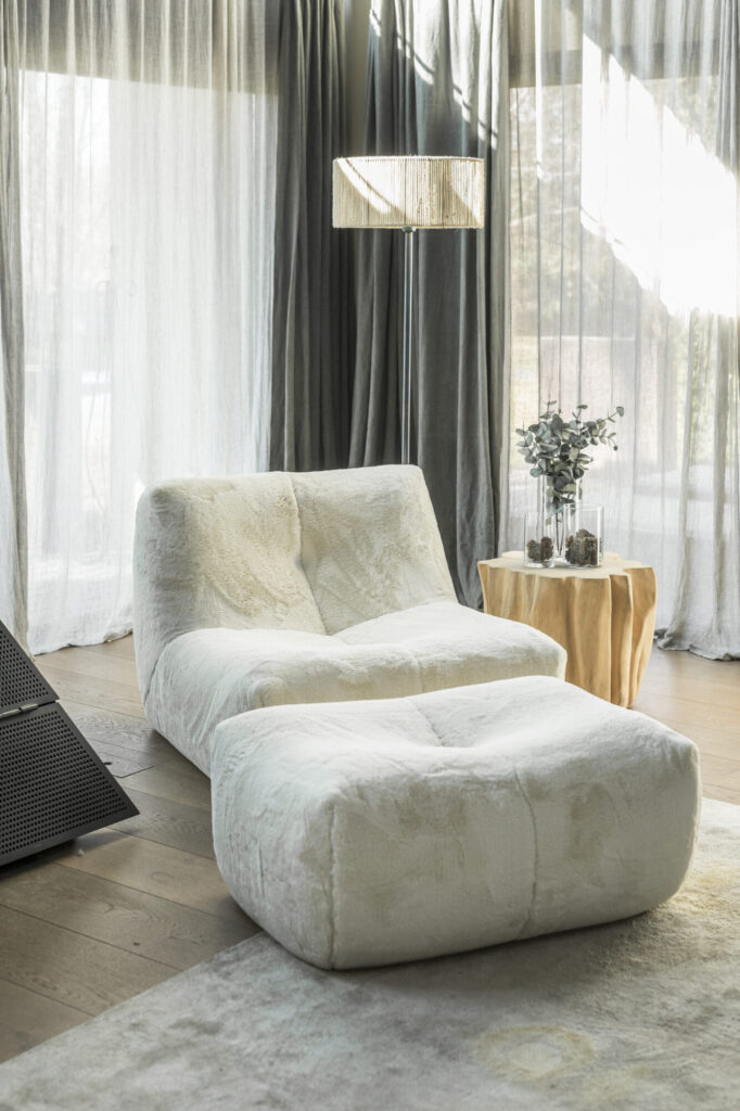 Le fauteuil Cozy S - 1 place CZ1 et le Pouf CZ23 assorti, tous deux en tissu blanc, sont placés à côté d'une petite table ronde avec un vase dans une pièce lumineuse dotée de rideaux à voiles allant du sol au plafond.