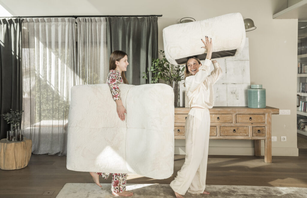 Deux personnes en pyjama jouent avec de grands coussins blancs moelleux à côté du Cozy S - 1 place CZ1 et du Pouf CZ23 dans un salon confortable avec des meubles en bois et de grandes fenêtres.